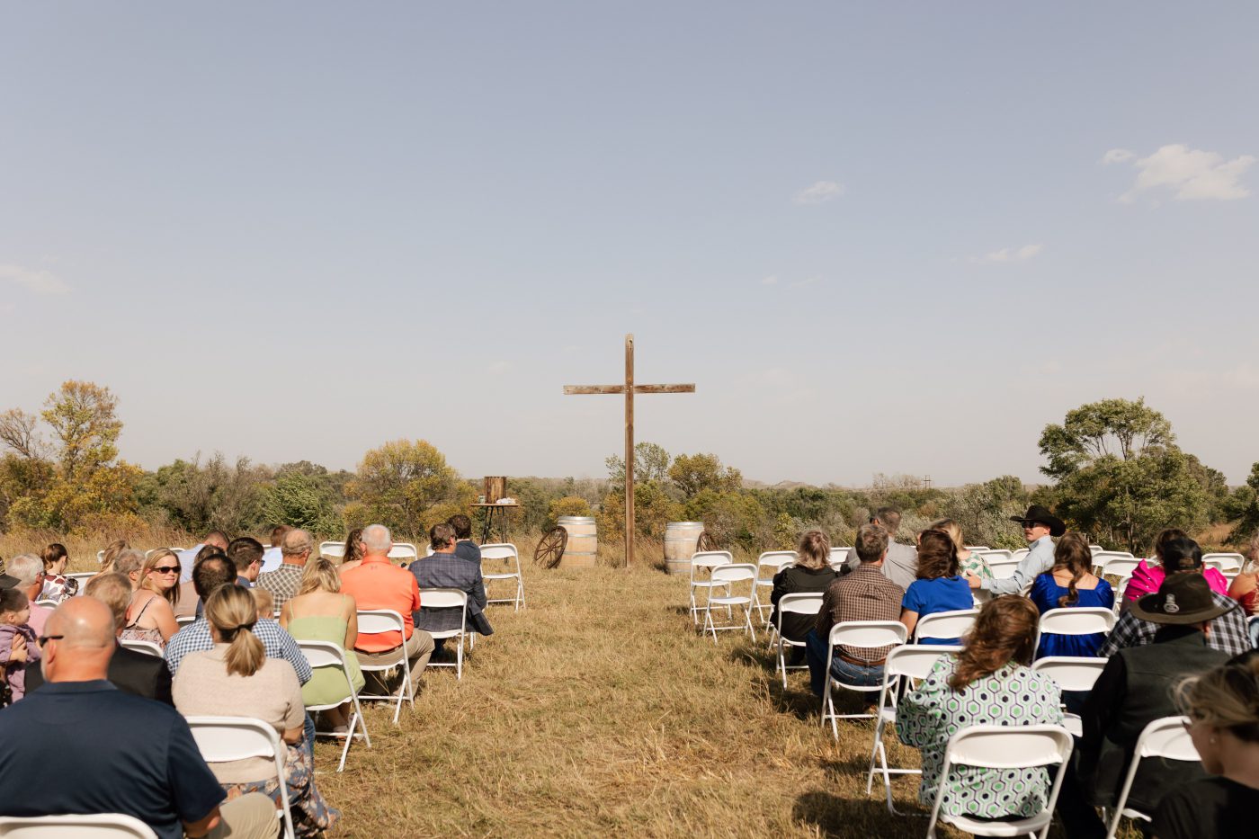A large wooden cross in an open field facing rows of wedding guests seated in white folding chairs