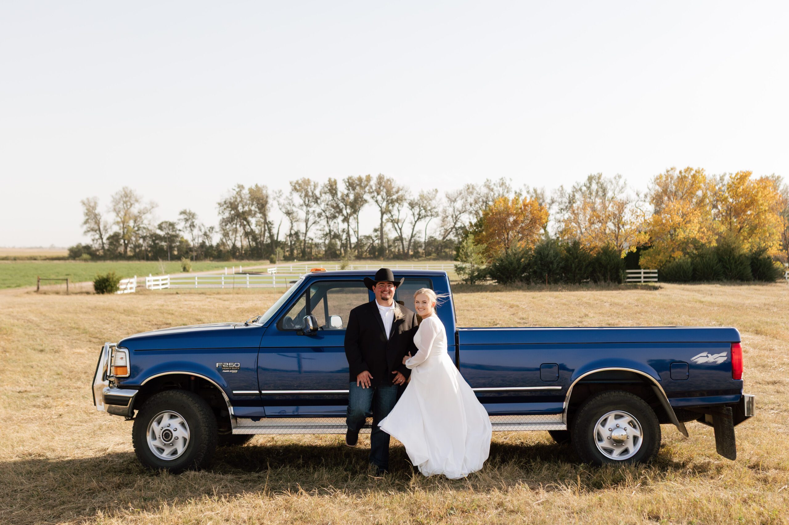 A bride and groom stand in front of a large blue pickup truck in the middle of a field in Central Nebraska